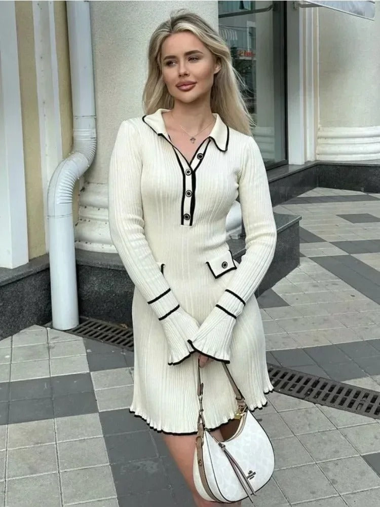 A lifestyle photo of an influencer standing on a European street, wearing a classy, white, ribbed dress with buttons, and a polo collar; she's holding a small purse in front, and looking to the side